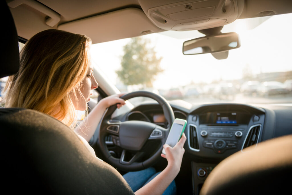 Young woman looking to her smartphone while driving car - rear view, sun shines through front window