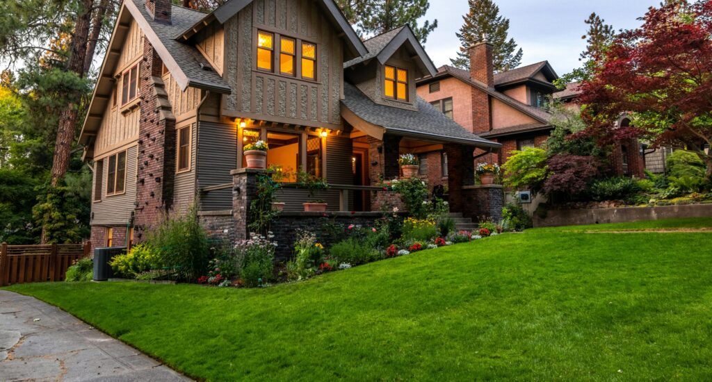 A warmly lit two‑story Craftsman-style house with a brick-and-wood exterior, a front porch lined with potted plants, and a colorful garden and trees surrounding the neatly trimmed lawn.
