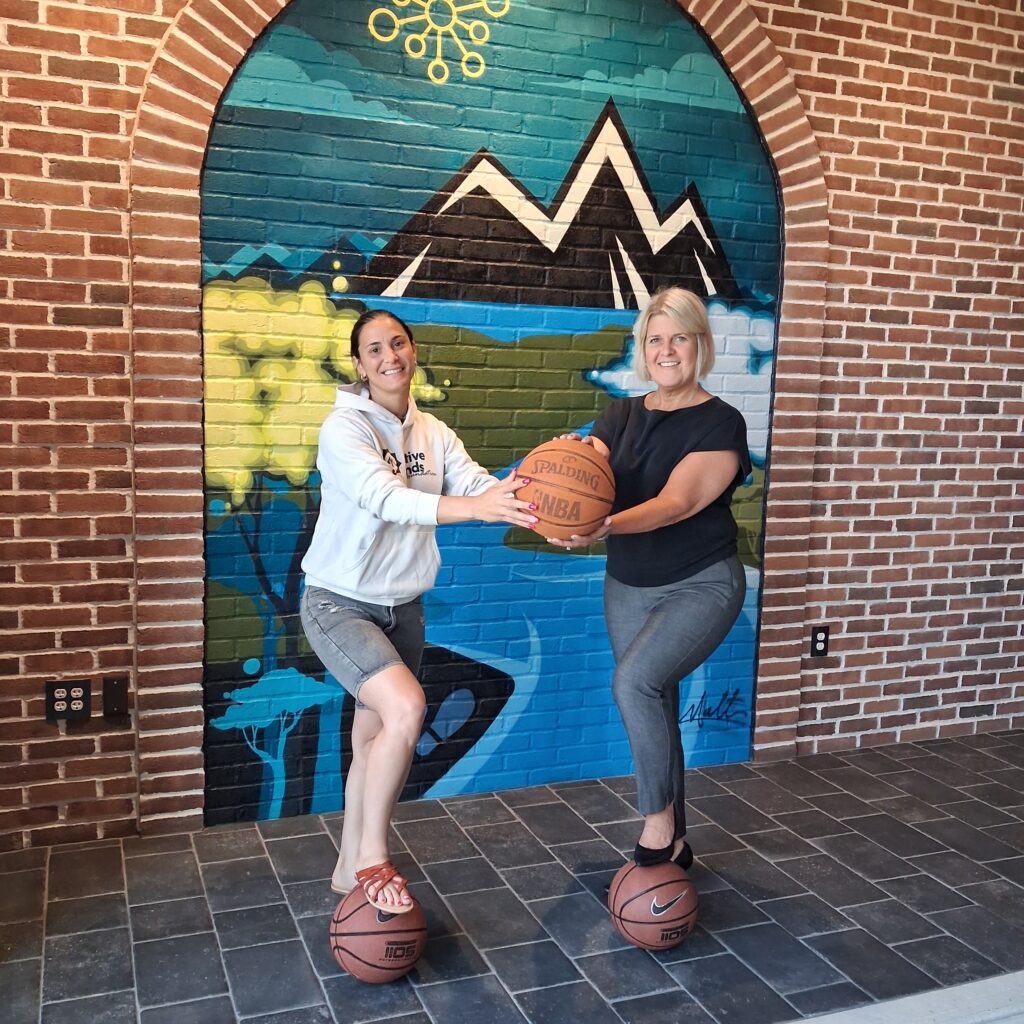Two individuals pose with a ceremonial check outside near a colorful mural and brick building.