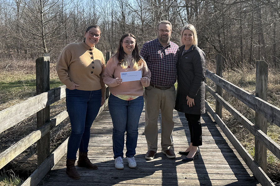 Four people standing on a wooden boardwalk in a natural outdoor setting, with one person holding an envelope during a donation presentation.