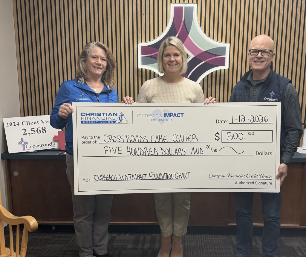 Three people hold a ceremonial $500 grant check for Crossroads Care Center in front of a branded wall display.