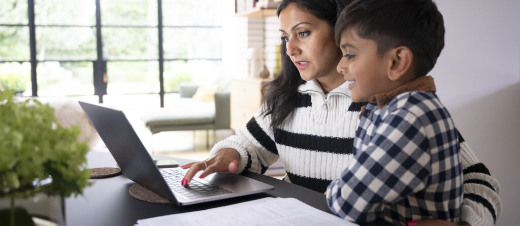 Parent reviewing financial resources on a laptop with a child at home, exploring ways to manage household expenses and access financial assistance programs.