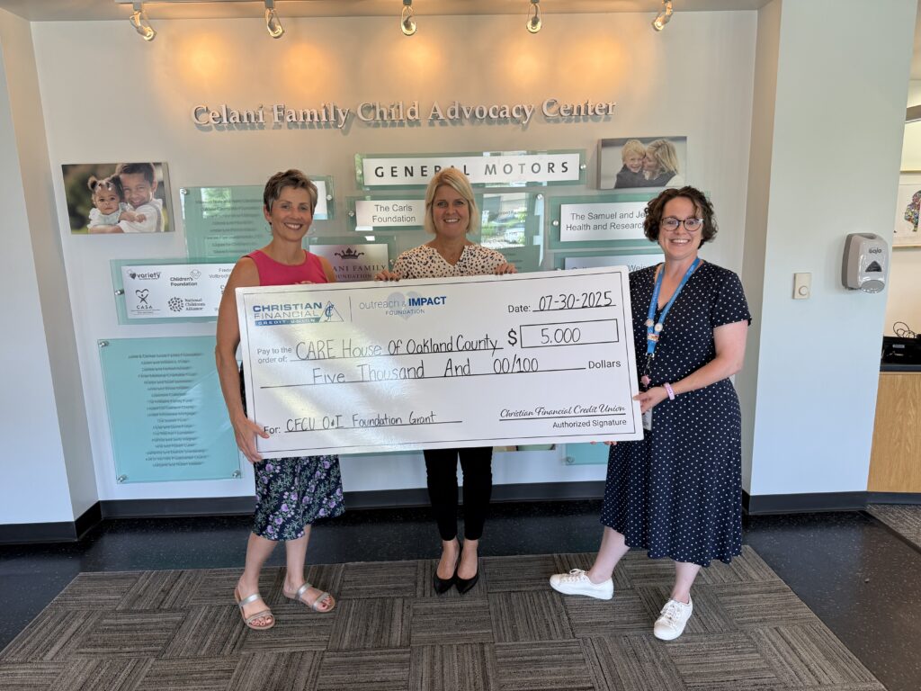 Three people standing indoors holding a ceremonial $5,000 grant check presented to CARE House of Oakland County.