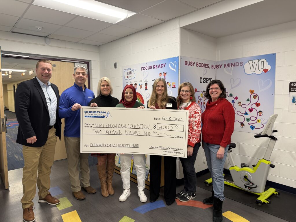 Seven people stand together holding a ceremonial $2,000 grant check for the Novi Educational Foundation inside a school learning space.